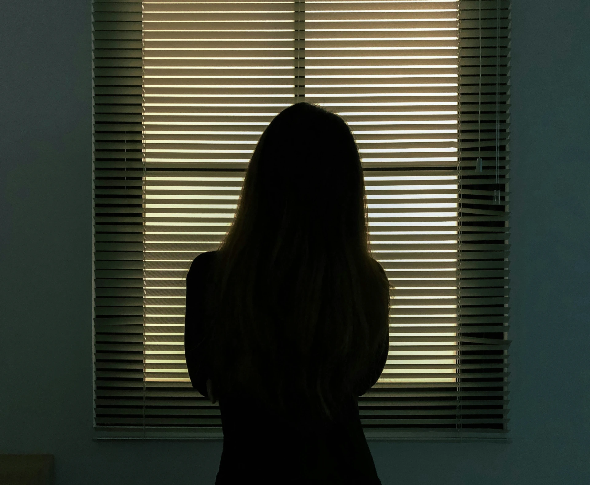A woman looking out a window slightly covered with blinds